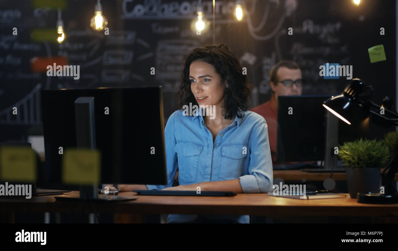 Bellissima femmina dipendente ufficio lavora alla sua scrivania su un Personal Computer. Sullo sfondo un collega in ufficio creativo. Foto Stock