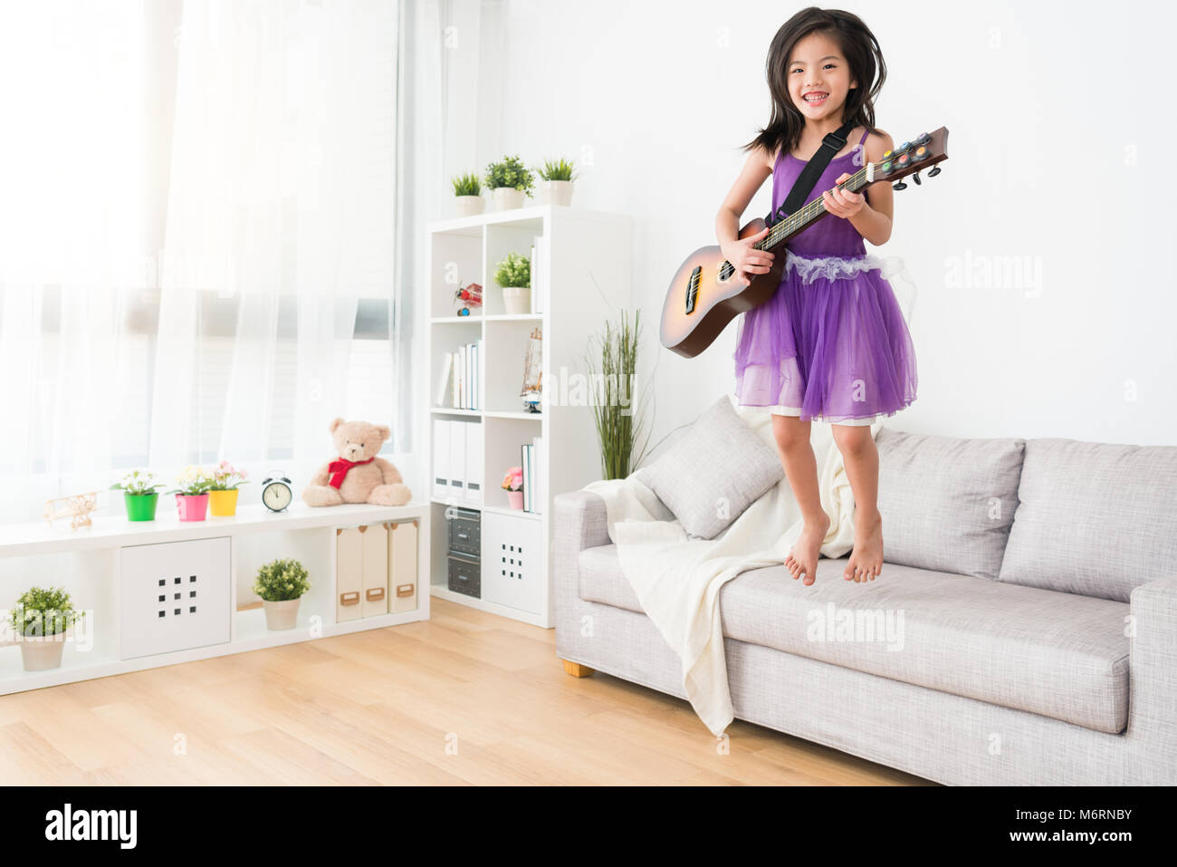 Energica ragazza in abito viola appoggiata dal divano. Essa detiene una chitarra e sorrisi con gioia. In una vista laterale. Sogna di diventare una femmina di bilanciere in Foto Stock