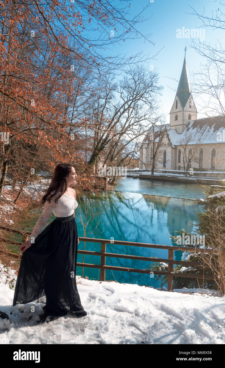 Elegantemente vestito giovane donna bruna, passeggiate attraverso la neve, sulla riva della molla di Blautopf, in una giornata di sole, a Blaubeuren, Germania. Foto Stock