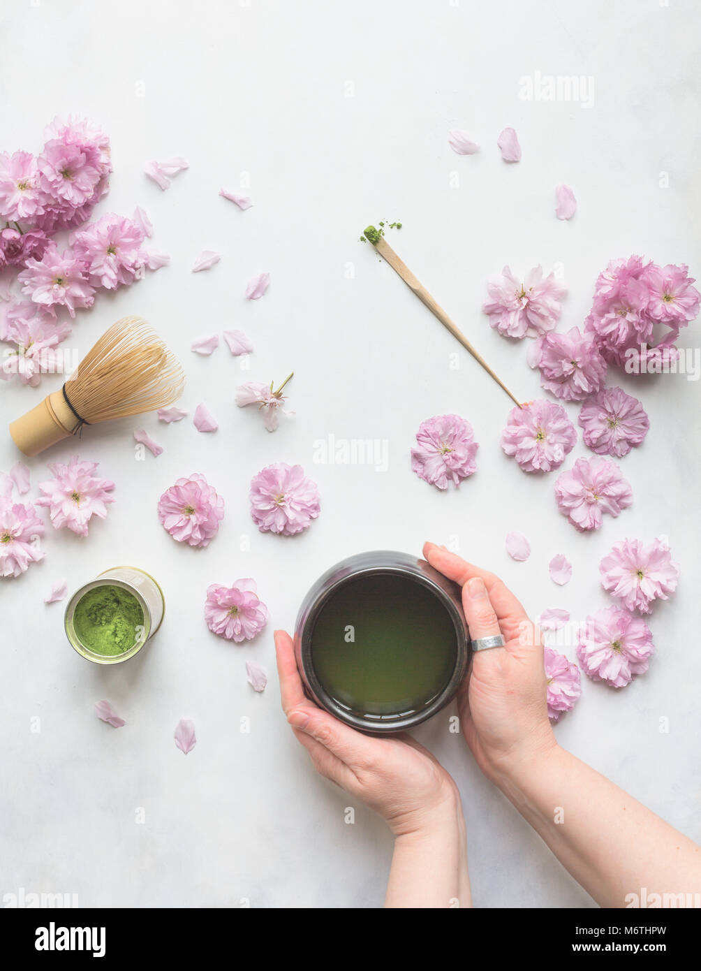 Vista dall'alto di due mani femminili tenendo un bicchiere di verde Matcha tè, con fiori di ciliegio, frusta e misurino. Foto Stock