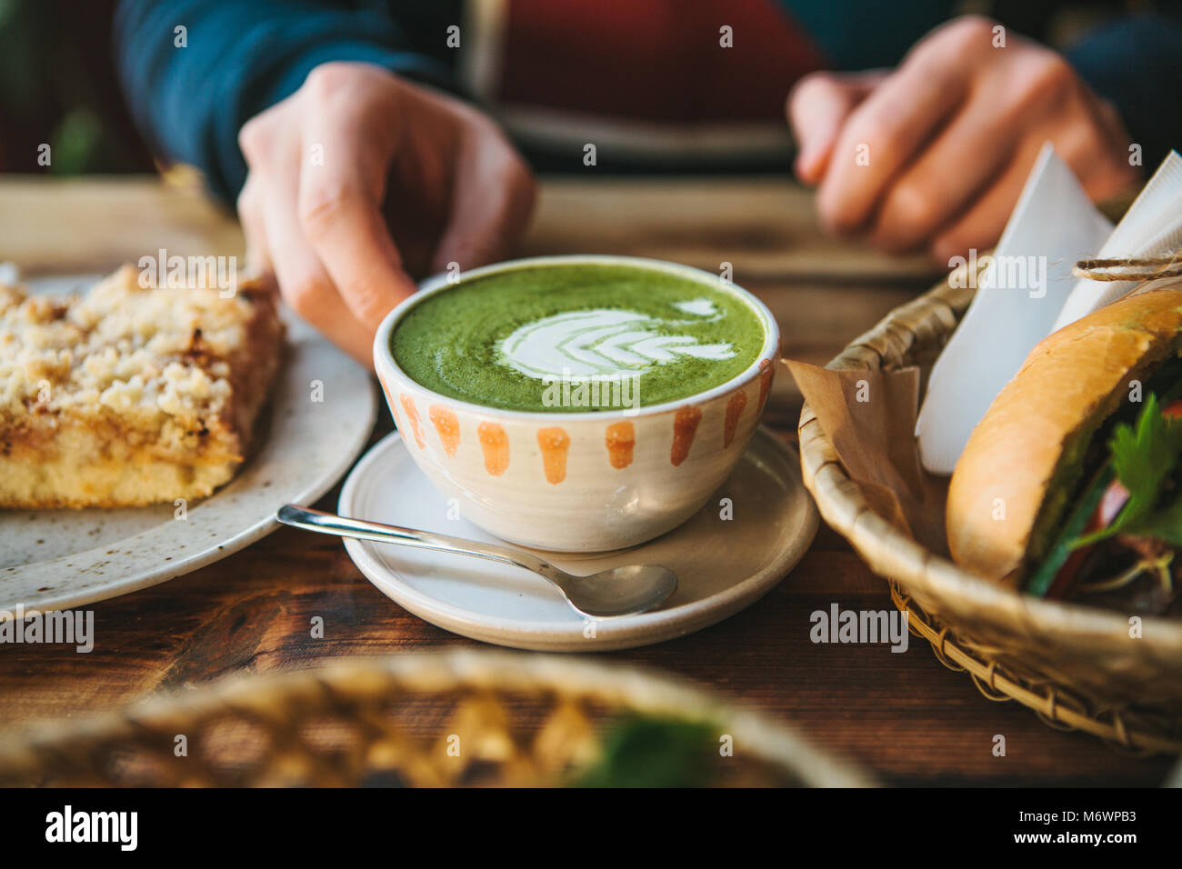 Close-up di man mano che tiene la tazza di tè verde con bellissimo motivo sotto forma di schiuma bianca accanto al dessert e sandwich Foto Stock