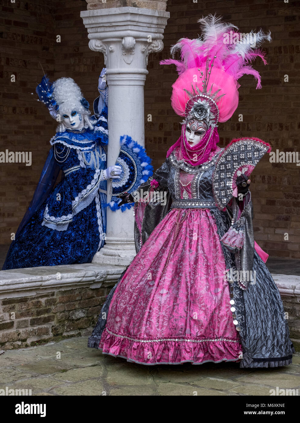 Donne mascherate in ornato blu e rosa costumi con ventole in piedi accanto a una colonna di pietra in un cortile interno durante il Carnevale di Venezia Foto Stock
