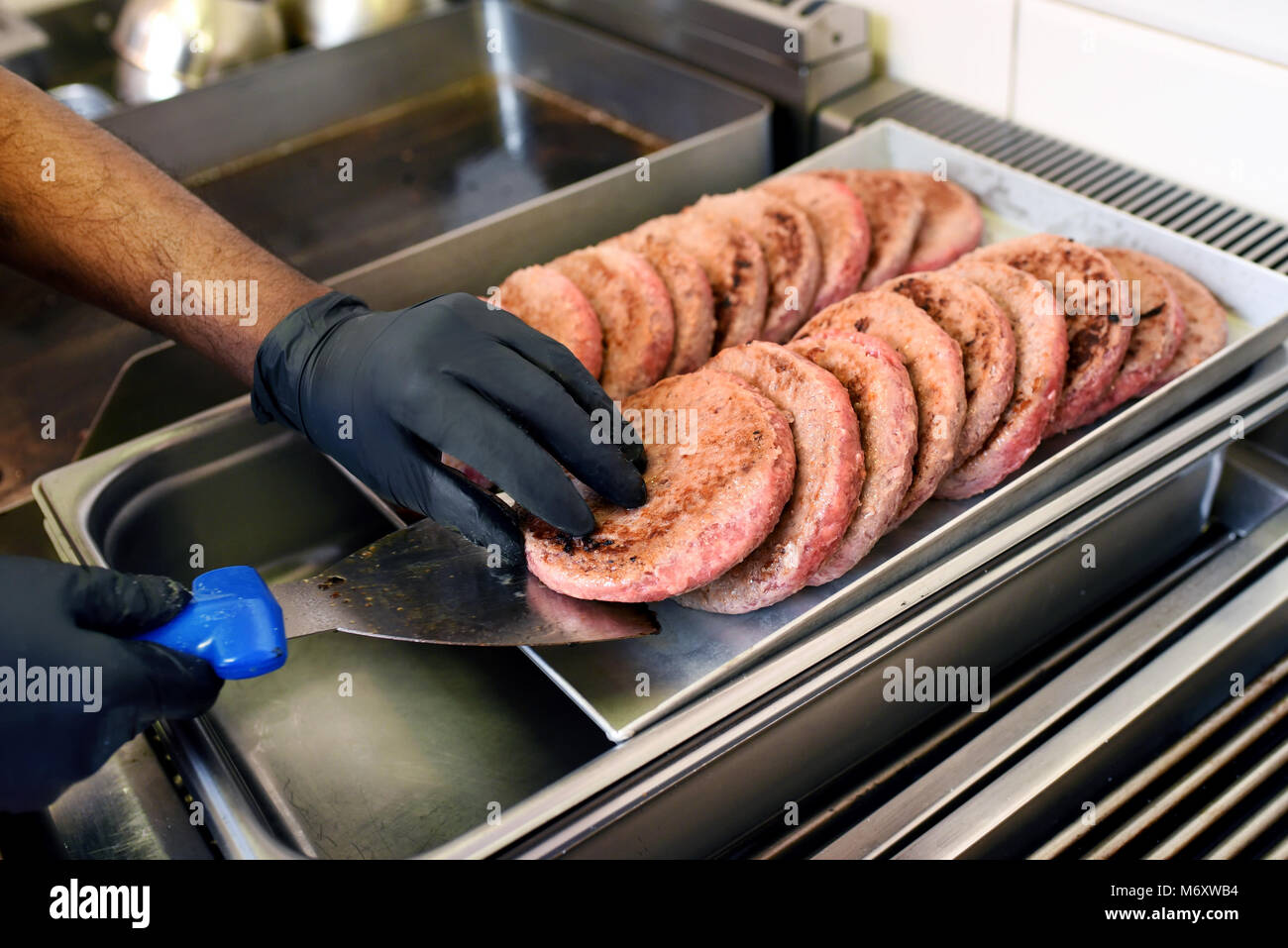 Lo Chef prendendo un pre-cotto hamburger di manzo patty fuori un vassoio di metallo utilizzando una spatola in un ristorante fast food Foto Stock