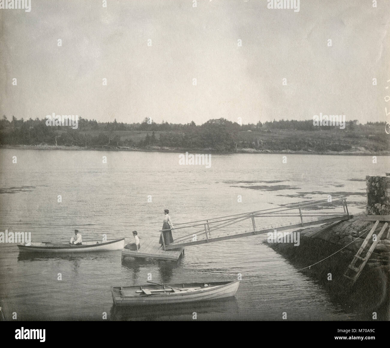 Antique circa 1905 fotografia, donne sul dock e uomo in canotto sul fiume Sasanoa. La posizione è in o vicino a Riggsville (ora Robinhood), Maine in Sagadahoc County, Stati Uniti d'America. Foto Stock