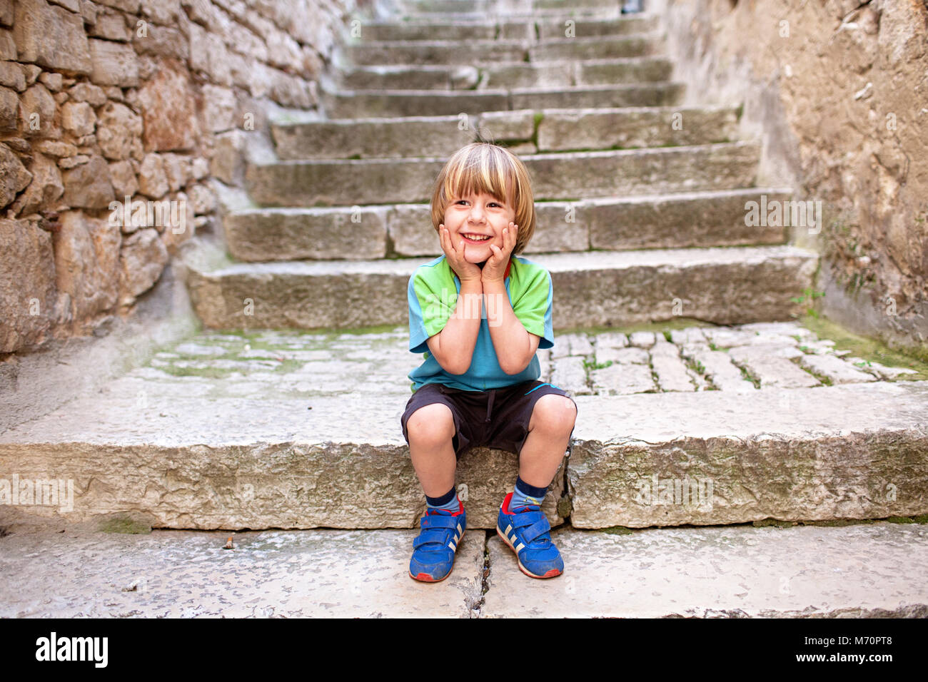 Carino biondo sorridente piccolo ragazzo seduto sulle vecchie scale di ciottoli con le mani sulle guance, nel centro storico di Rovigno Foto Stock