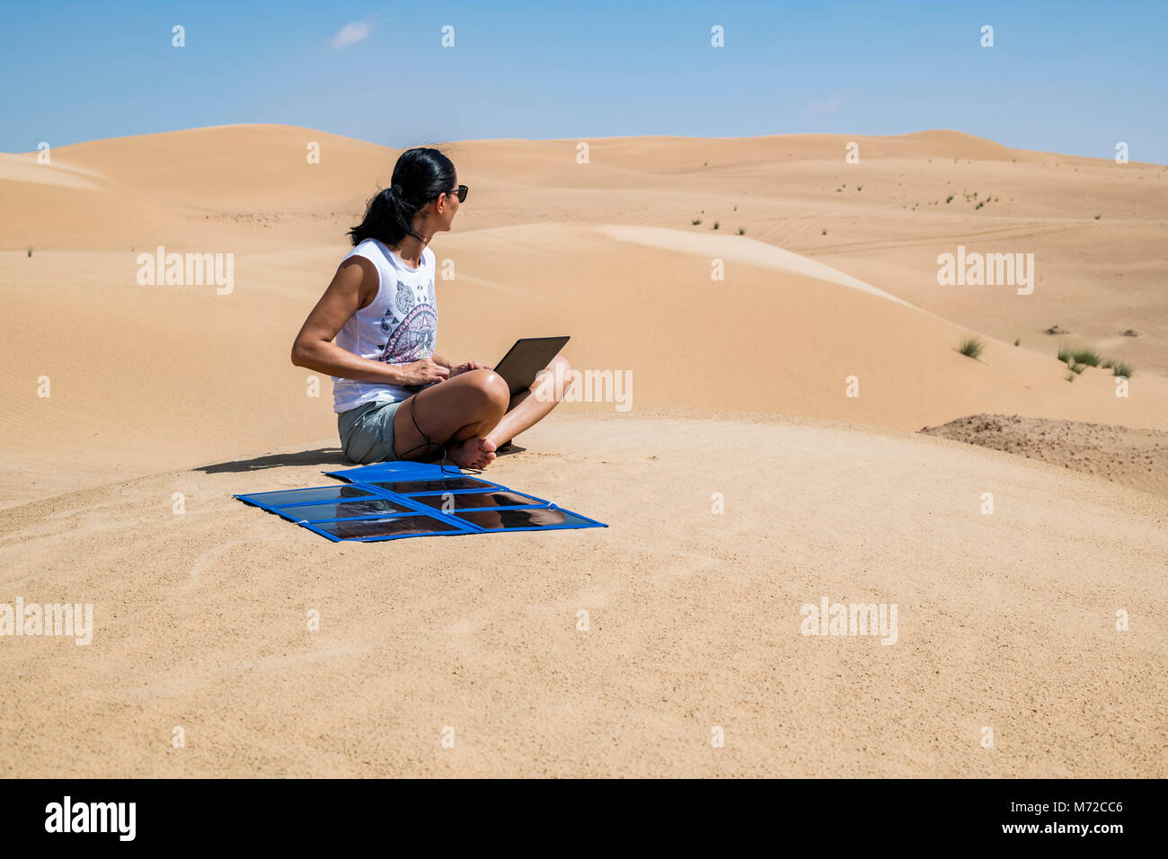 Una donna turista utilizza il suo laptop tra le dune con un pannello solare portatile, autosufficiente e sostenibile mentre lavora in remoto in una posizione remota Foto Stock