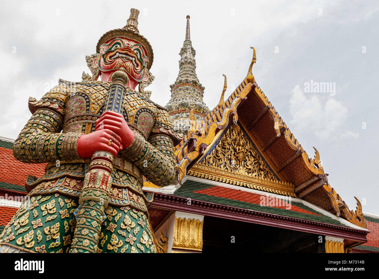 Vista dal basso della statua di una guardia al Grand Palace di Bangkok, con il frontone di un tempio in background Foto Stock