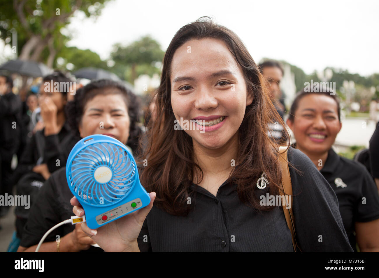 Ragazza sorridente con un ventilatore portatile, e le donne in attesa in linea per assistere al Royal cremazione dell amato re in Bangkok. Tutto nero di medicazione Foto Stock