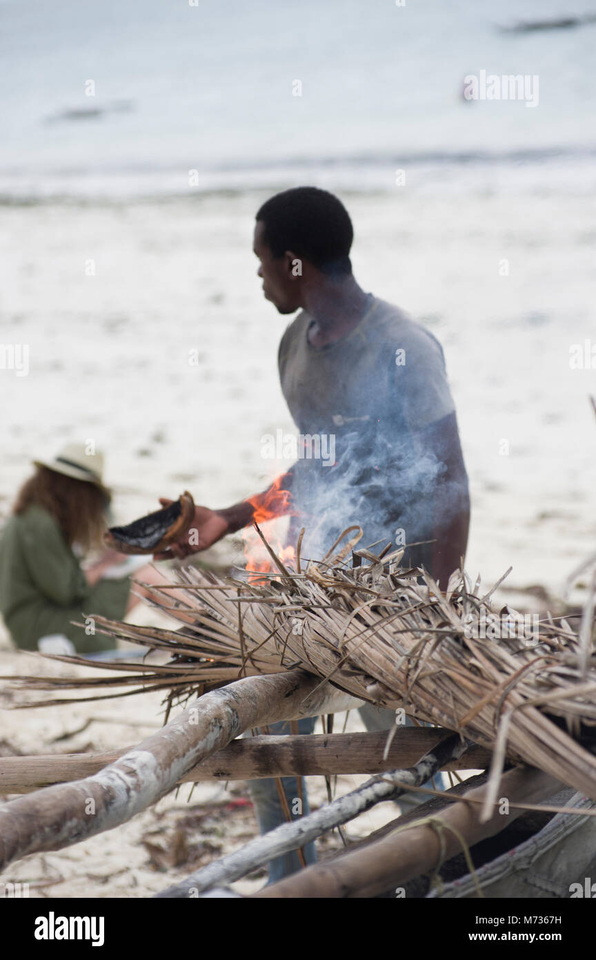 Fisherman utilizzando la masterizzazione di fronde di palma per rimuovere il muschio e verde crescite erbosa dal lato della sua nave da pesca in Jambiani Zanzibar Tanzania Foto Stock
