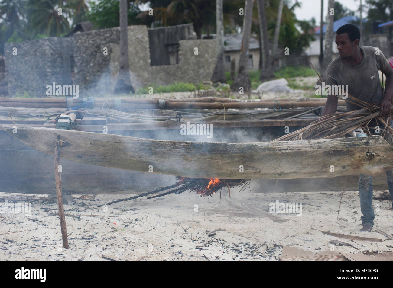 Fisherman utilizzando la masterizzazione di fronde di palma per rimuovere il muschio e verde crescite erbosa dal lato della sua nave da pesca in Jambiani Zanzibar Tanzania Foto Stock