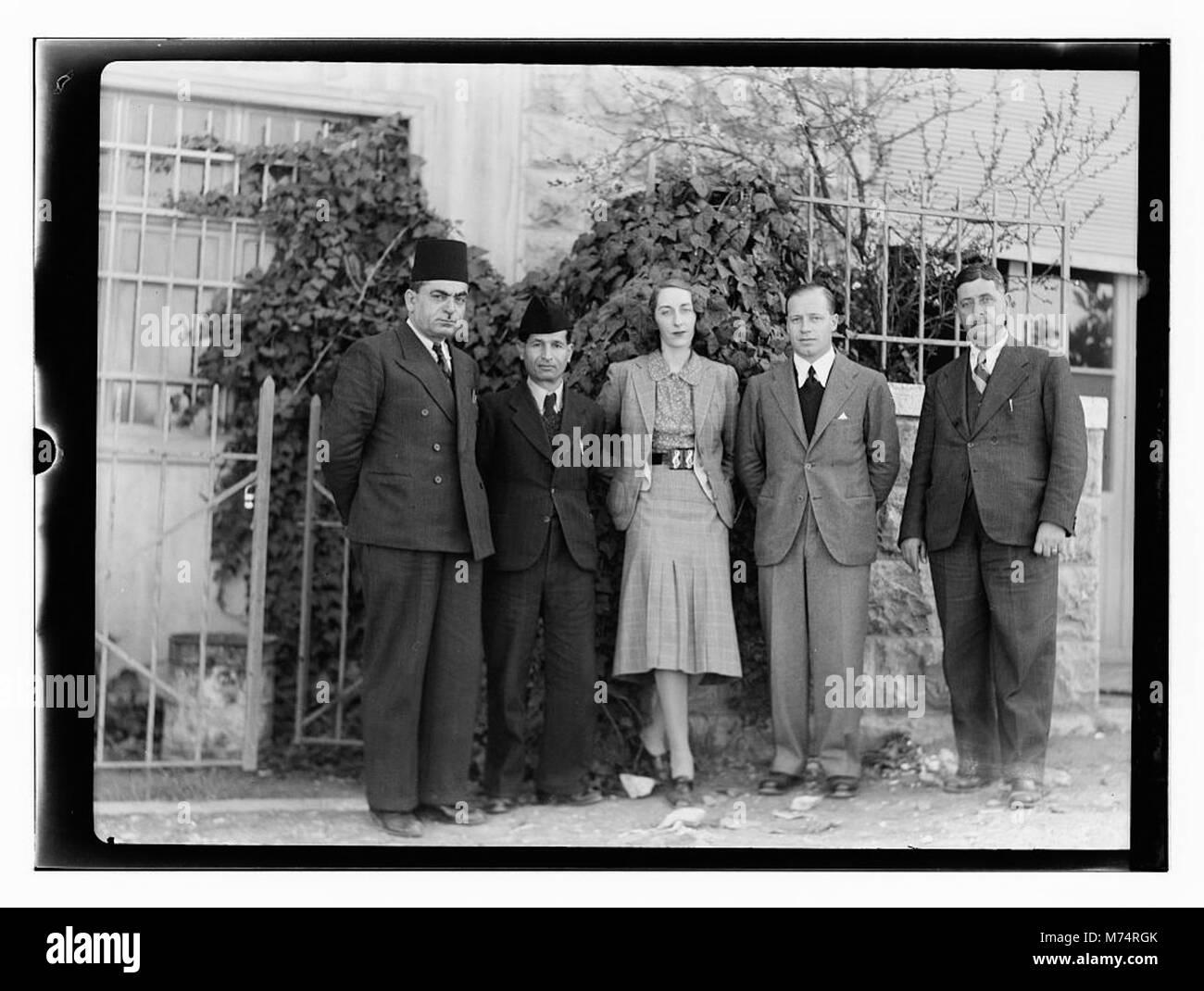 Questo ritratto di gruppo presenta una donna in piedi con quattro uomini di fronte a un edificio, offrendo uno sguardo sulla vita sociale e sui ruoli di genere durante la fine del XIX secolo o l'inizio del XX secolo. Foto Stock