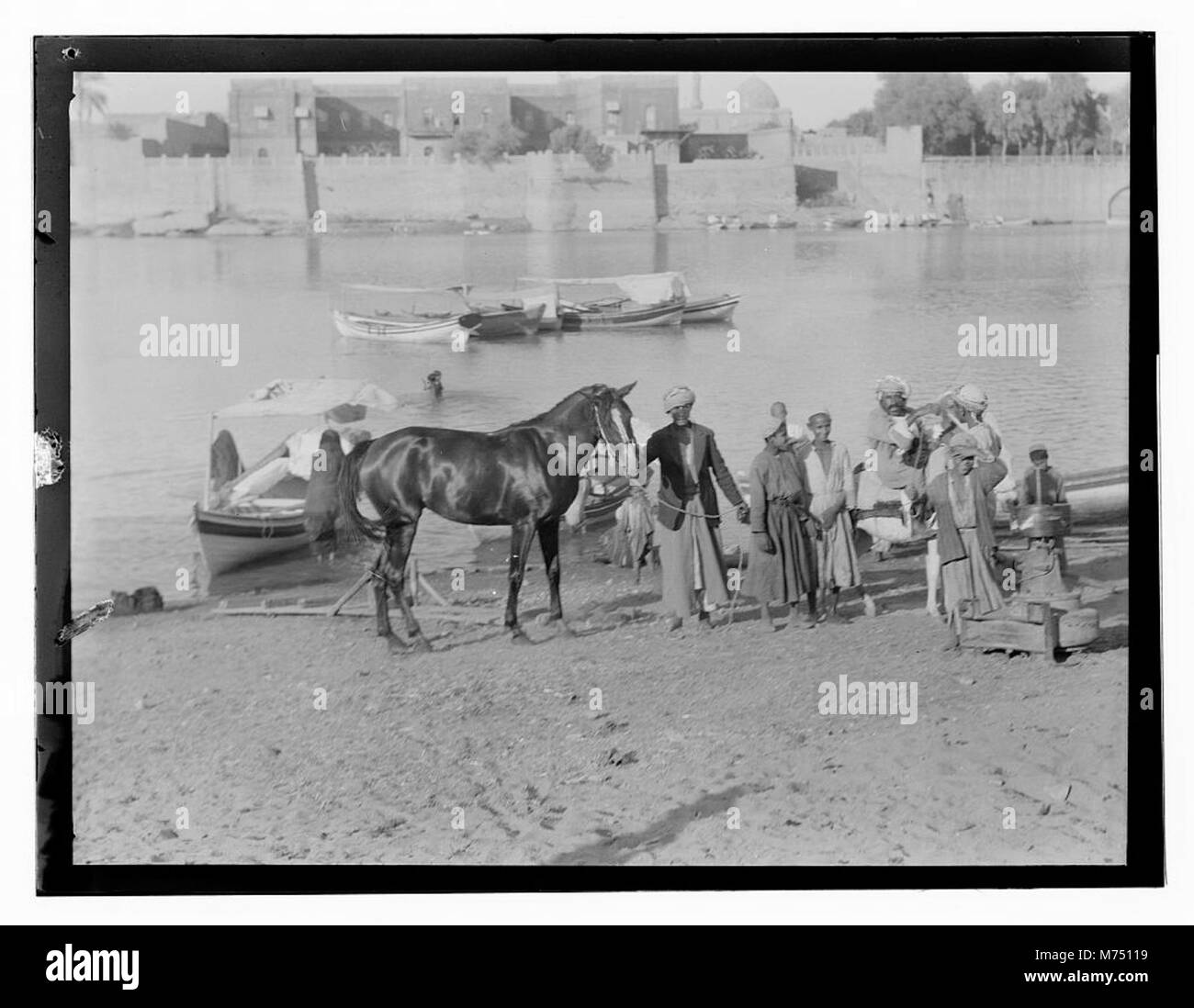 Questa fotografia mostra uomini e un cavallo vicino a un fiume, con una città murata sullo sfondo. L'immagine cattura una scena rurale in Iraq, evidenziando l'interazione tra le persone e il loro ambiente. Foto Stock