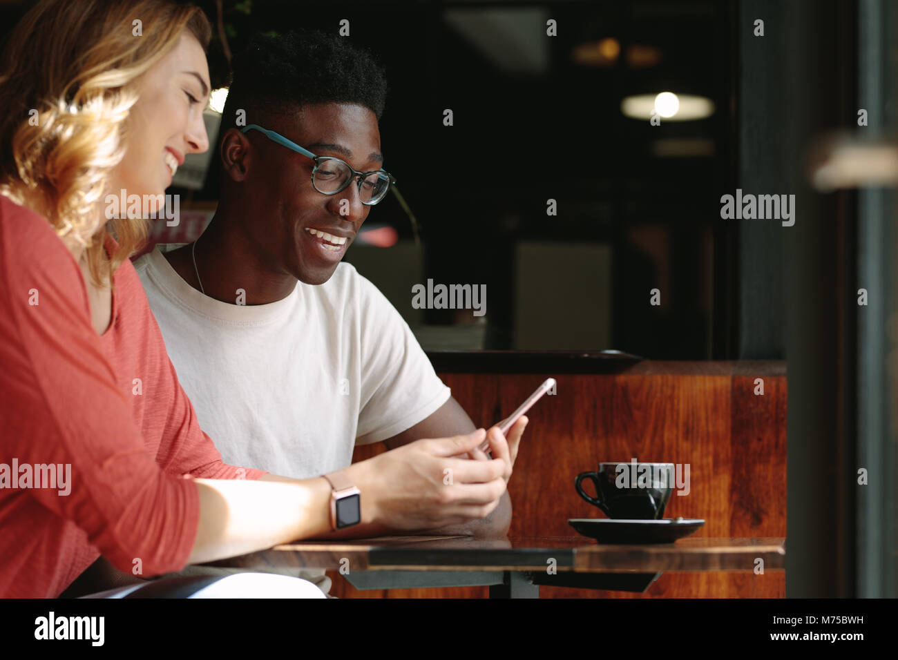 L uomo e la donna seduta al tavolo da caffè guardando un telefono cellulare. Coppia felice in un coffee shop utilizzando il telefono cellulare con una tazza di caffè sul tavolo. Foto Stock