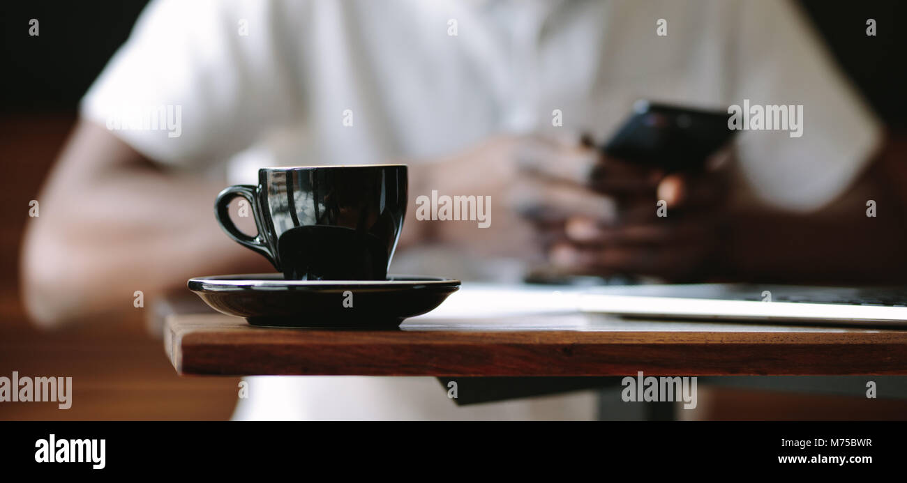 Primo piano di una tazza di caffè posto su di una tavola in una caffetteria. Ritagliato colpo di uomo seduto in un bar con un nero lucido tazza da caffè con piattino su Foto Stock