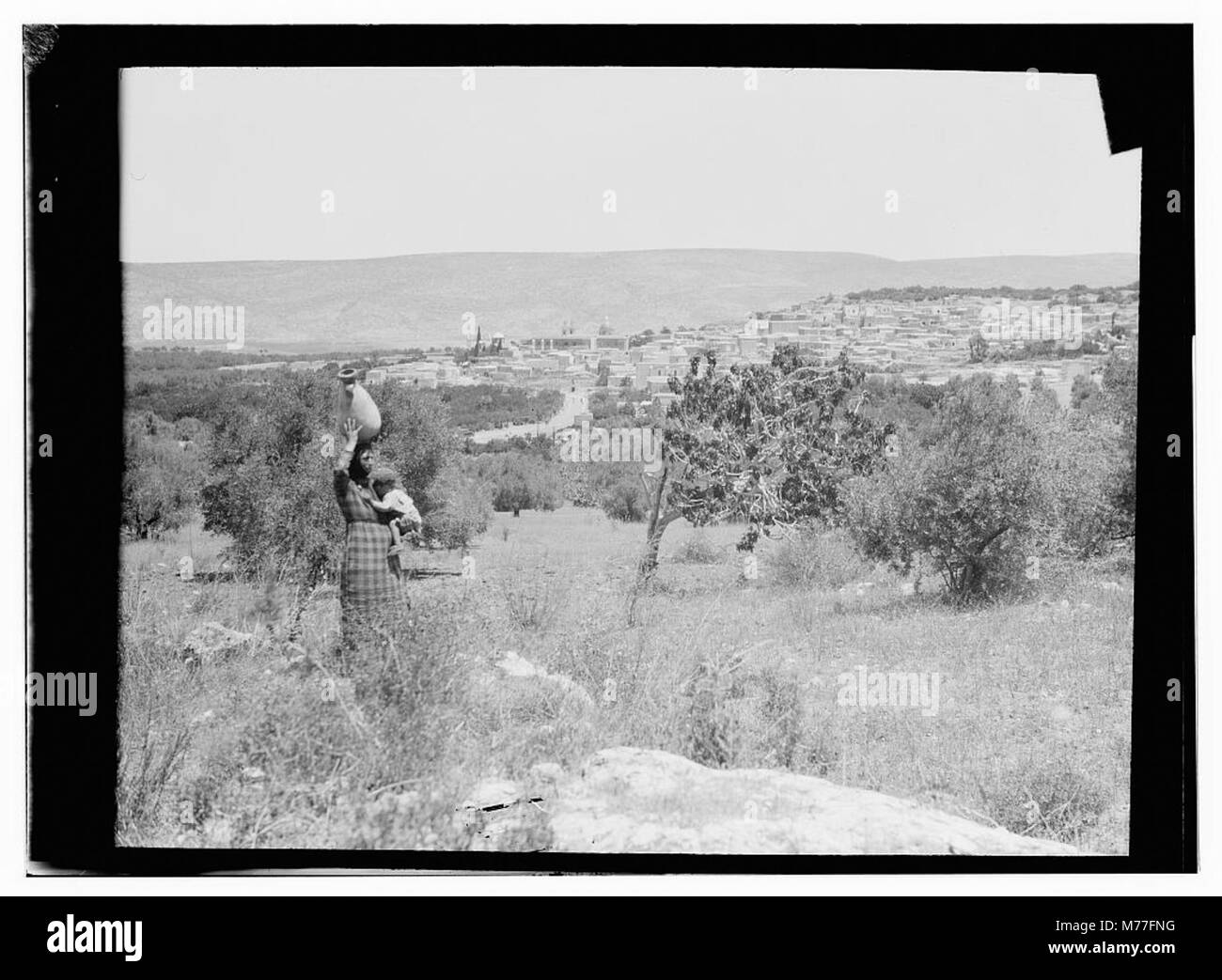Si vede una donna in piedi con un barattolo equilibrato sulla testa, con Nazareth visibile in lontananza. La scena riflette i costumi tradizionali e la vita quotidiana di questa regione, catturati all'inizio del XX secolo. Foto Stock