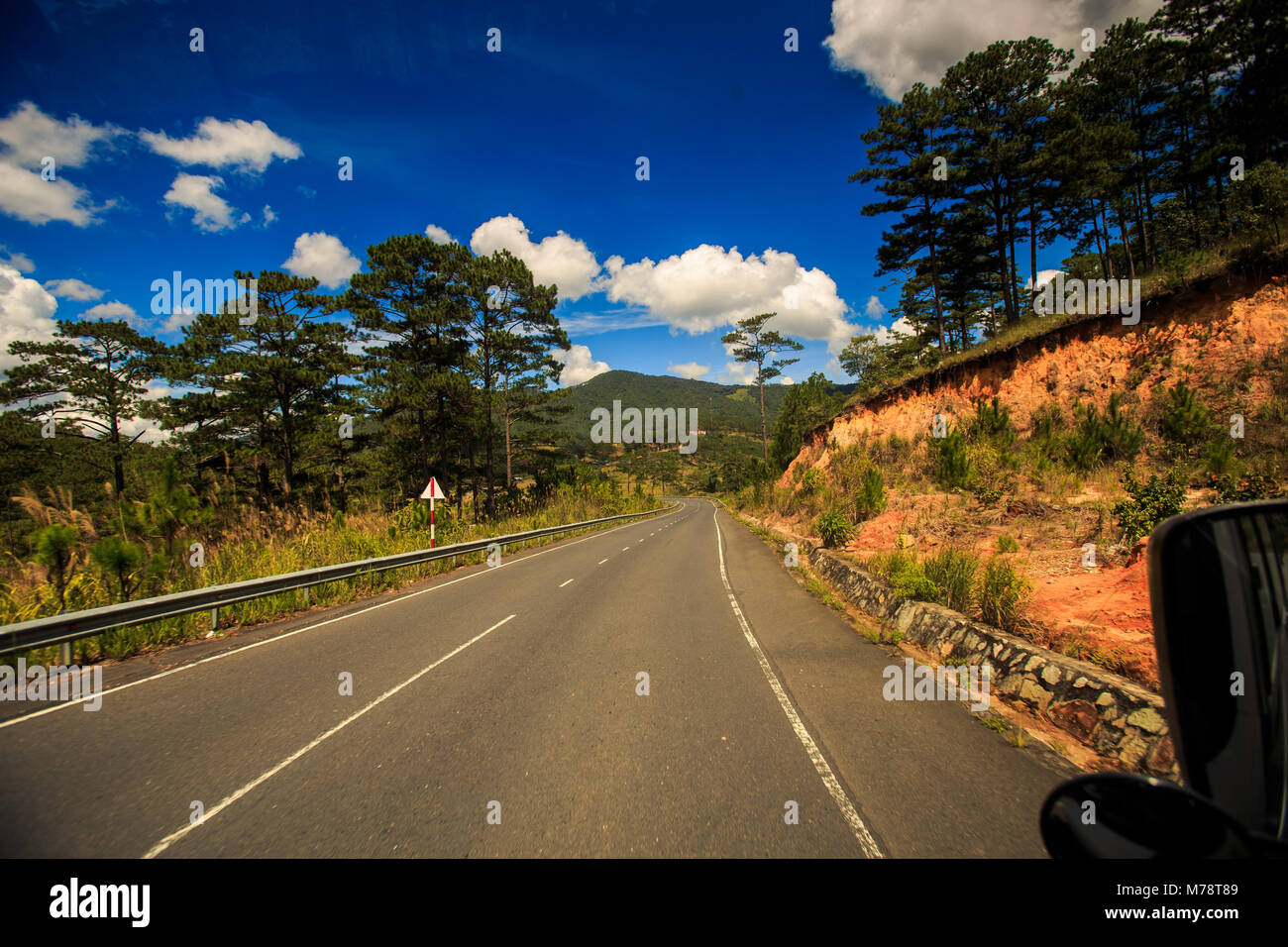Sunny autostrada lungo la collina di marrone e piante tropicali e driver specchio laterale contro nuvoloso cielo blu al di fuori della finestra di bus Foto Stock
