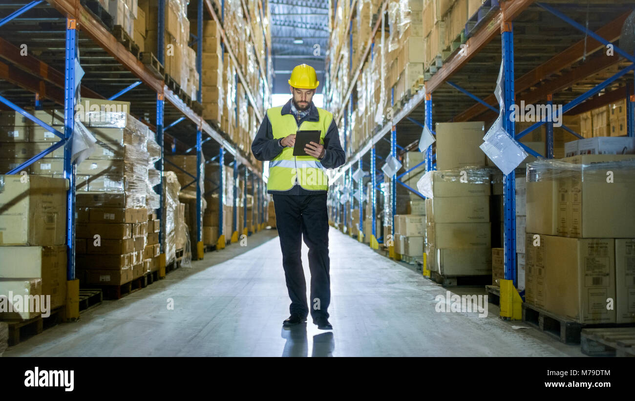 Revisore dei conti INDOSSARE ELMETTO con computer Tablet conta merce in magazzino. Egli cammina attraverso le righe dei rack di stoccaggio con la merce. Foto Stock