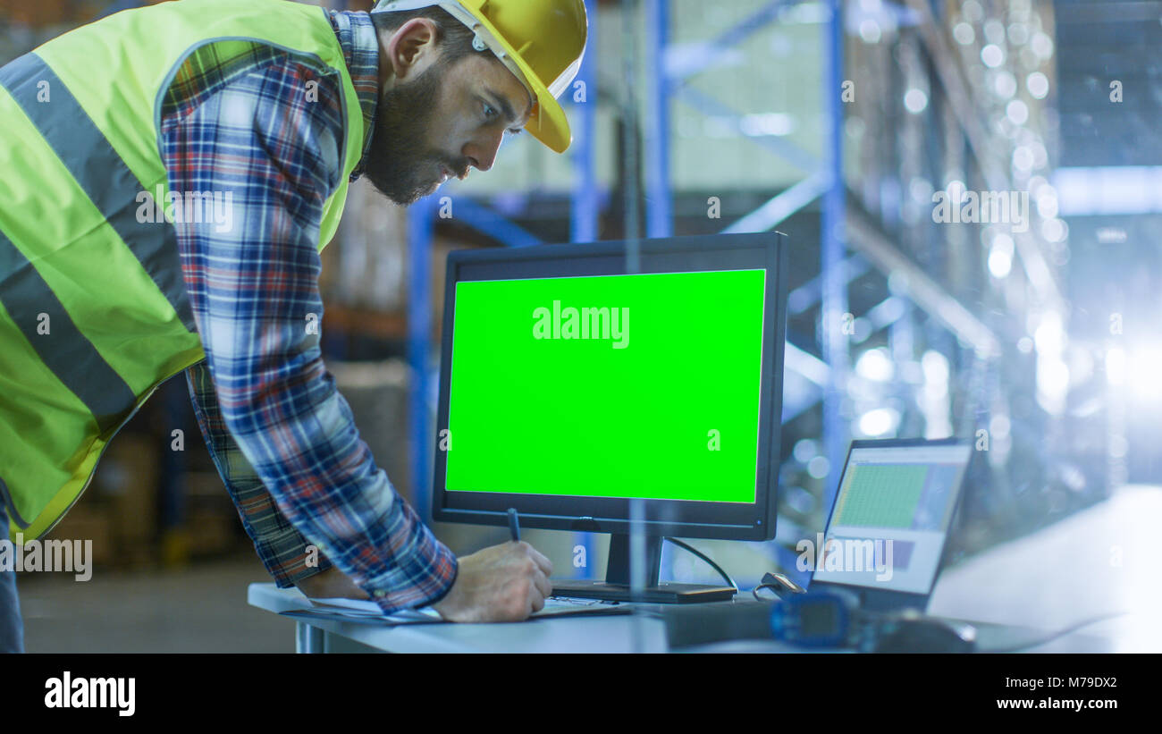 Ispettore maschio riempie i fogli di calcolo sul Suo Personal Computer con schermo verde modello grande per mockup. Egli è nel grande magazzino con righe Foto Stock