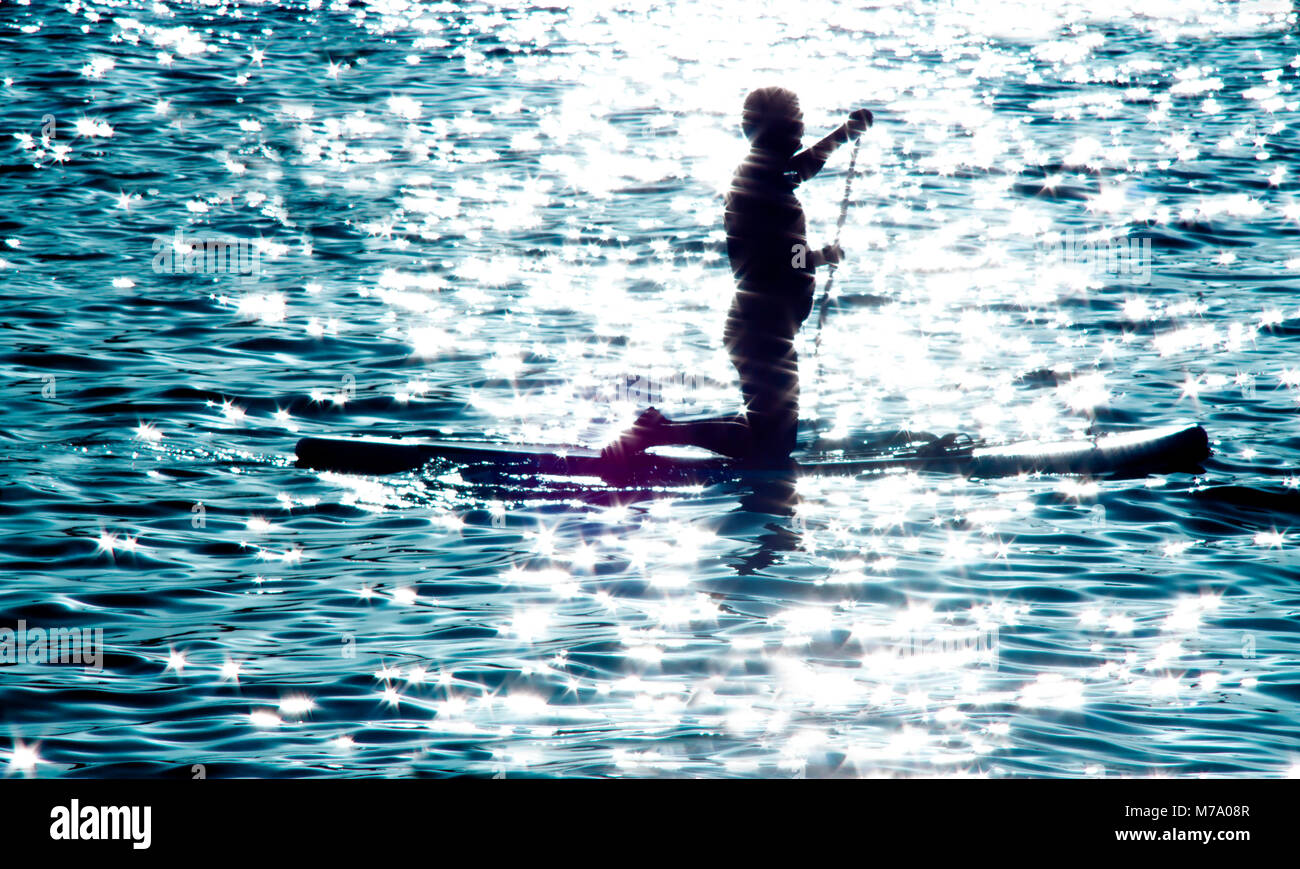 Sfocata silhouette di un uomo inginocchiato sulla scheda di racchetta e canottaggio al chiaro di luna illuminata mare, facendo sport di acqua ricreazione per il corpo e per la mente Foto Stock