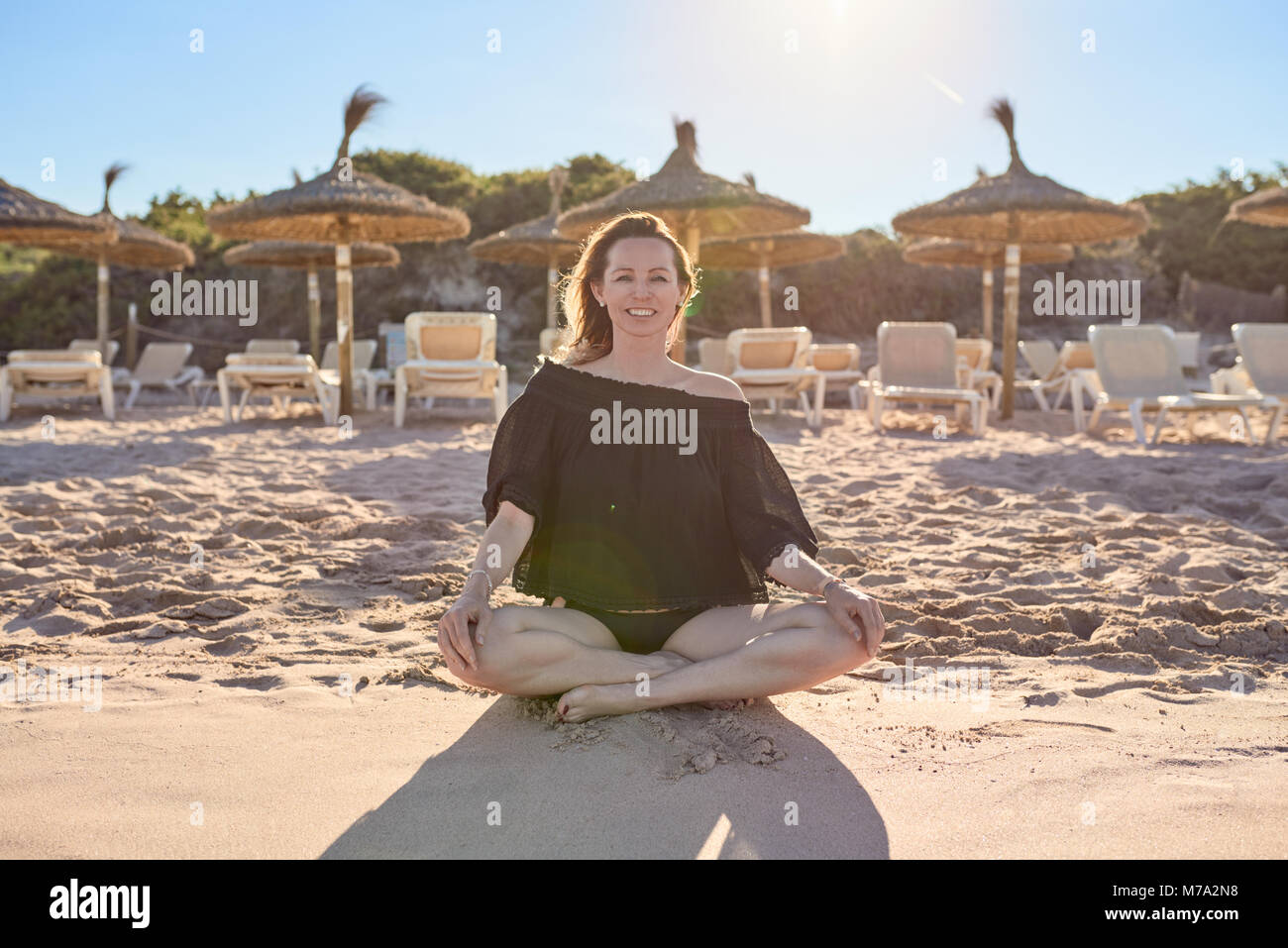 Salutare sorridendo a piedi nudi donna seduta con le gambe incrociate sulla spiaggia come si prepara a meditare sullo sfondo di un villaggio sulla spiaggia ombrelloni retroilluminati da t Foto Stock