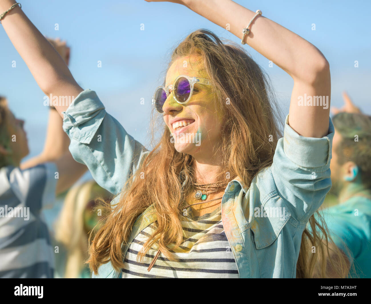 Close-up ritratto di una giovane e bella ragazza con occhiali da sole in piedi in mezzo alla folla di persone celebrare Holi festival. La gente di gettare polvere colorata Foto Stock