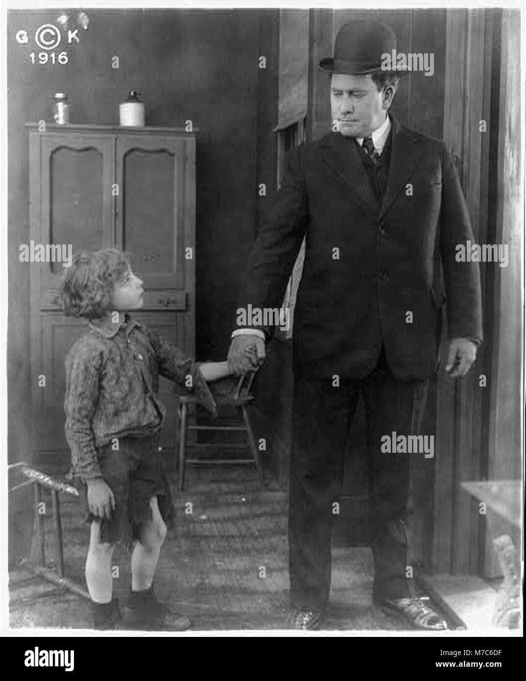 Un'immagine tratta da un film con un uomo che indossa un cappello derby, che tiene la mano di un ragazzo vestito con pantaloni tagliati, probabilmente da un film classico. Foto Stock