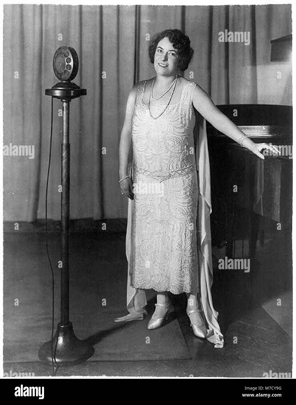 Un ritratto a figura intera di Frances Alda, in piedi tra un pianoforte e i microfoni radio. L'immagine riflette la sua carriera nelle arti dello spettacolo, catturandola come musicista o cantante professionista. Foto Stock