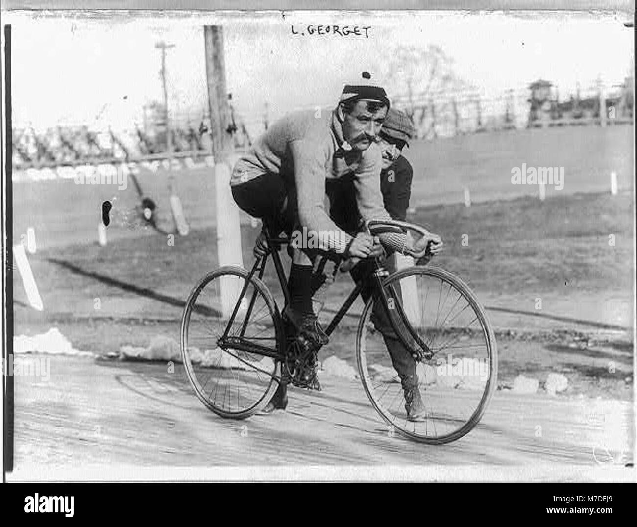 Questo ritratto a figura intera mostra Leon Georget seduto su una bicicletta, rivolto verso destra, mentre un altro uomo tiene la bicicletta, catturando un momento di cultura ciclistica e interazione personale. Foto Stock