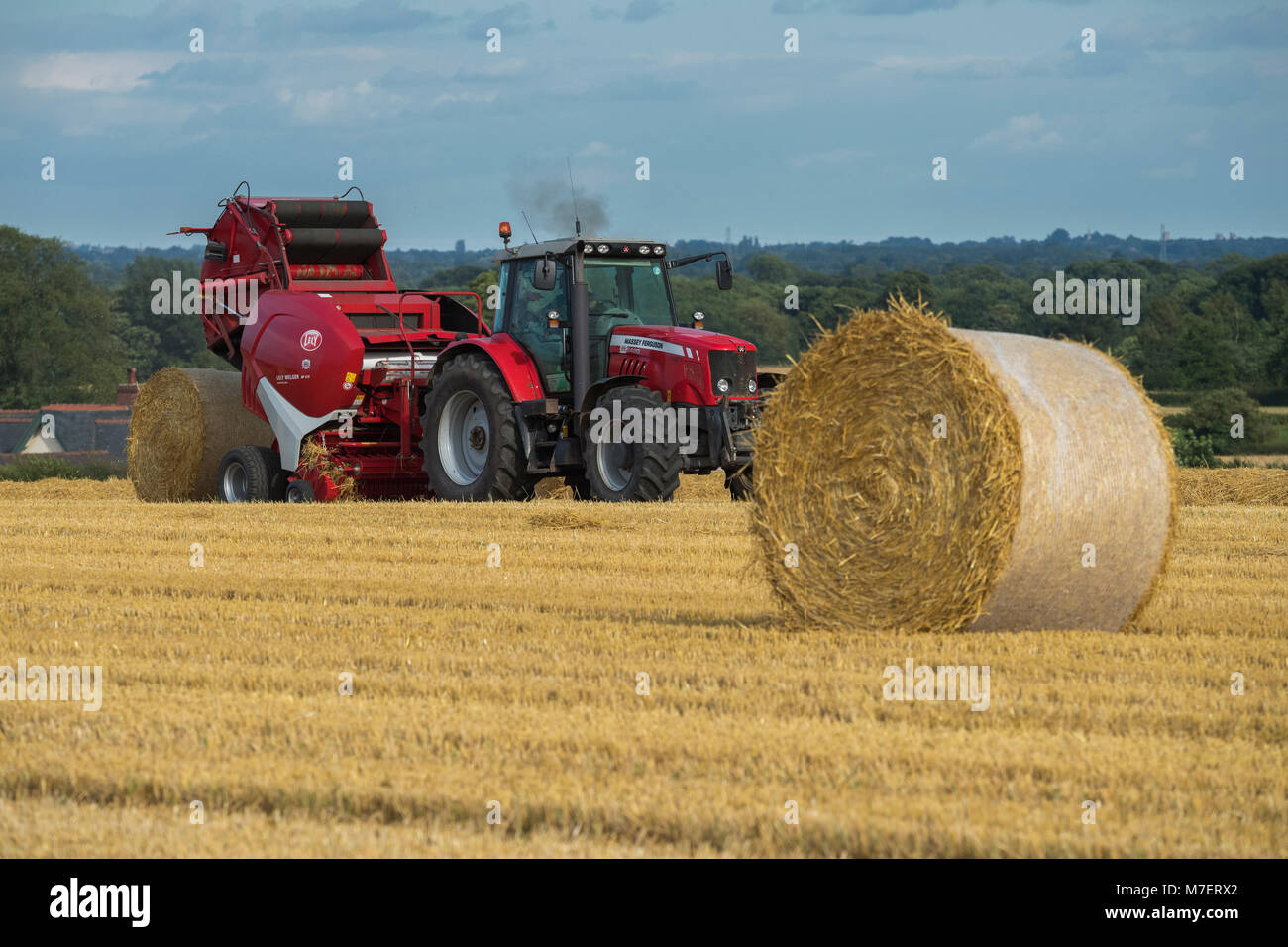 Le balle di paglia in una fattoria campo, agricoltore lavora e unità trattore rosso tirando la rotopressa (grande balla appena rilasciato) - Whixley, North Yorkshire, Inghilterra, Regno Unito. Foto Stock