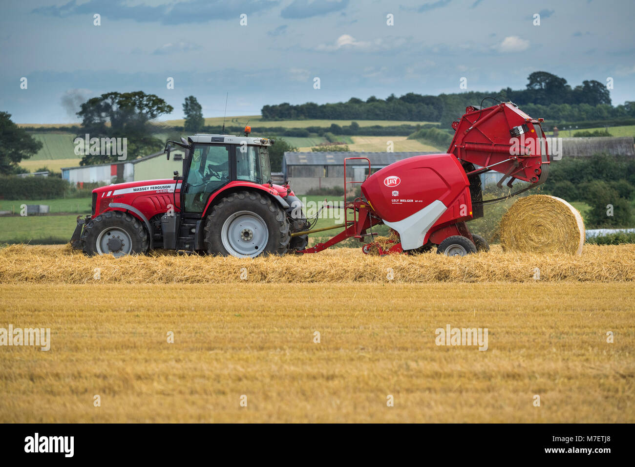 Le balle di paglia in una fattoria campo, agricoltore lavora e unità trattore rosso tirando la rotopressa (grande balla appena rilasciato) - Whixley, North Yorkshire, Inghilterra, Regno Unito. Foto Stock