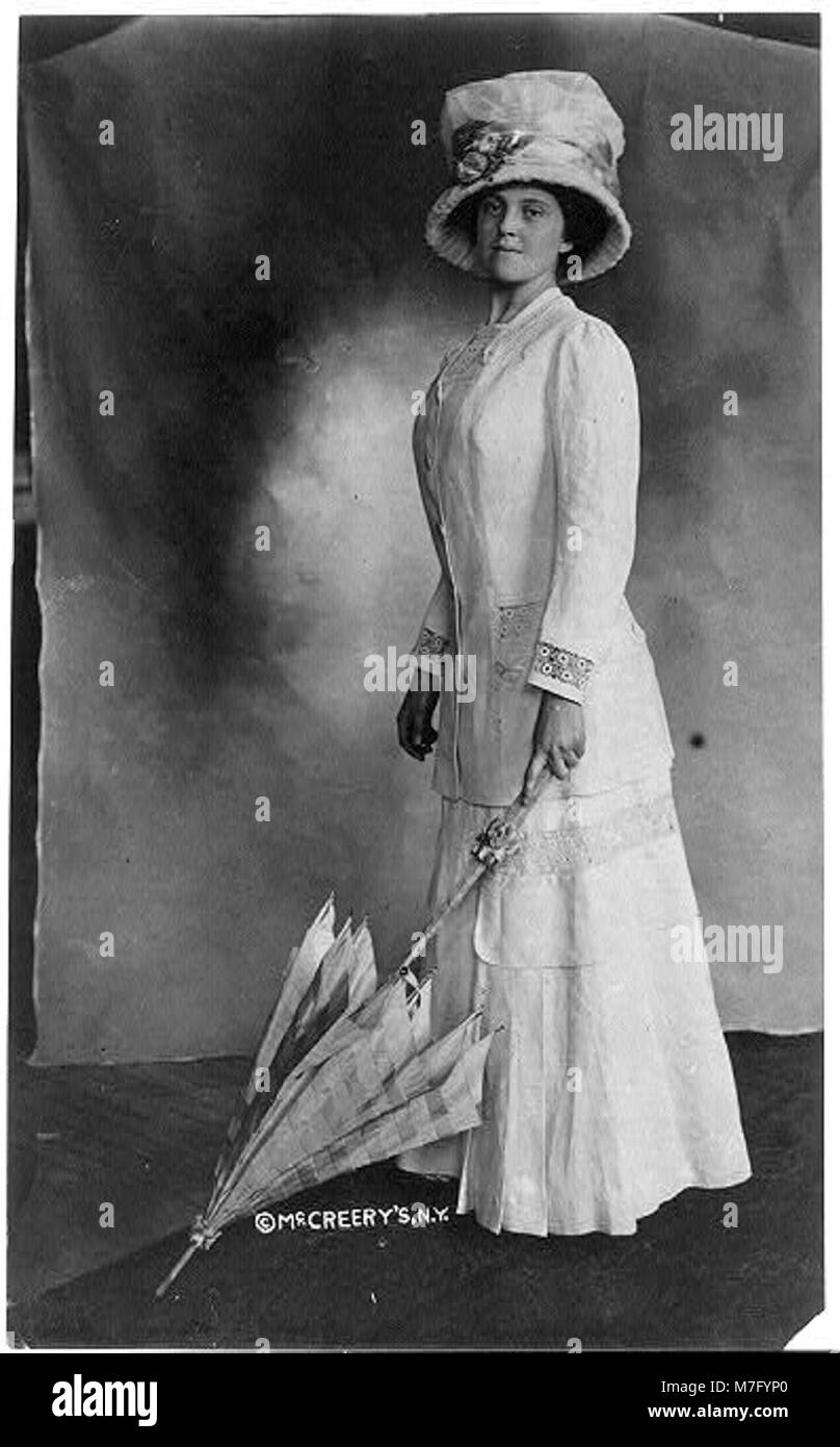 Una donna vestita con un abito a tutta lunghezza, un cappello e con in mano un ombrello, con la punta appoggiata sul pavimento. Questa fotografia mette in risalto la moda e lo stile dei primi anni del XX secolo. Foto Stock