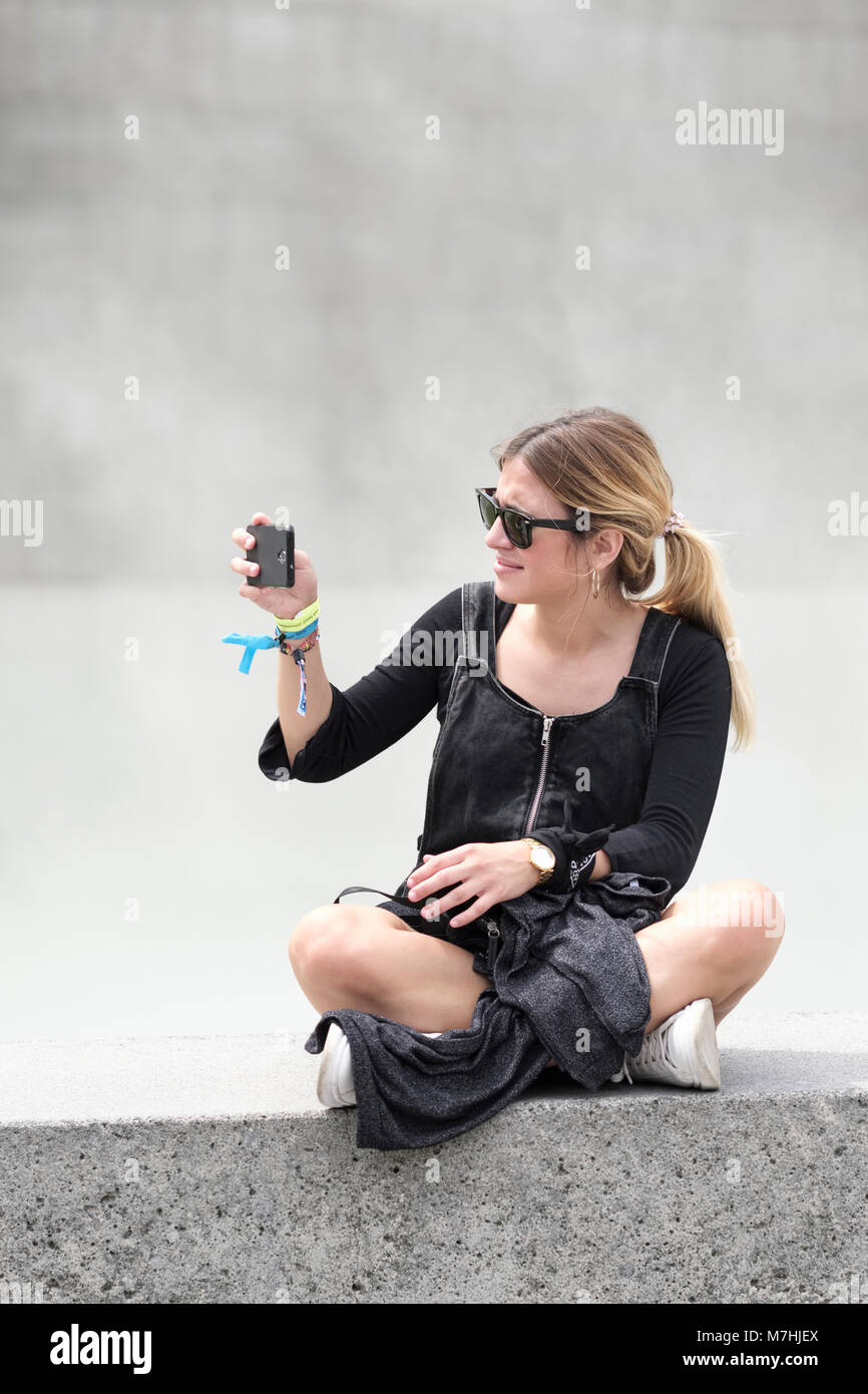 Bionda e giovane signora con la coda di cavallo, seduto sul muro con il telefono cellulare, prendendo un selfie, Bilbao Vizcaya, Pais Vasco, Spagna, Foto Stock