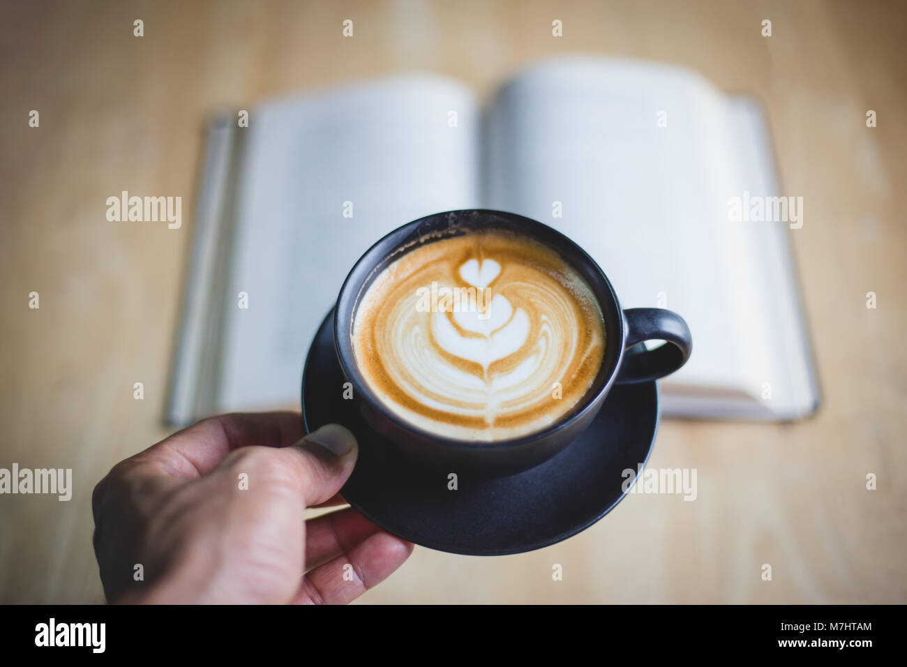 Mano che regge una tazza di latte con caffè e sfocato libro sul tavolo Foto Stock