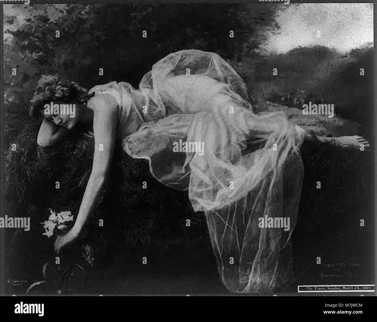 Questa immagine ritrae una giovane donna in un abito grazioso, sdraiata sulla riva di un corpo d'acqua. Sta toccando delicatamente un fiore, catturato a tutta lunghezza. Foto Stock