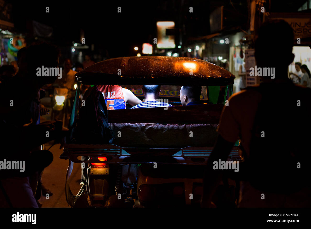 Due maschi di passeggeri e conducente sono illuminati in un sidecar moto taxi durante la guida lungo un affollato notte tempo street di Coron Town, Busuanga ho Foto Stock