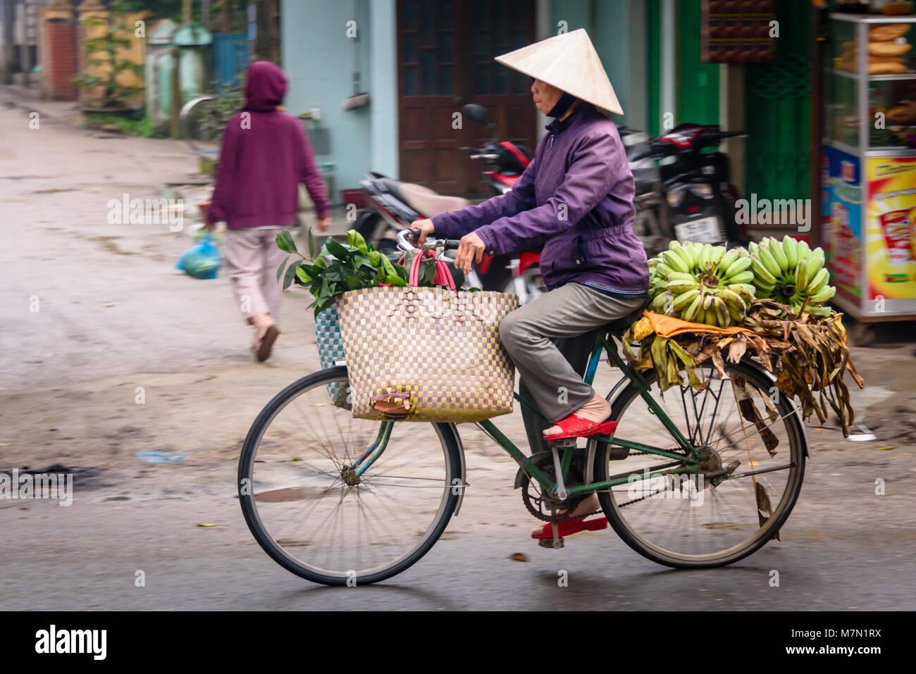 Una donna vietnamita che indossa un tradizionale cappello conico di bambù fa un giro in bicicletta con banane e frutta in vendita. Foto Stock