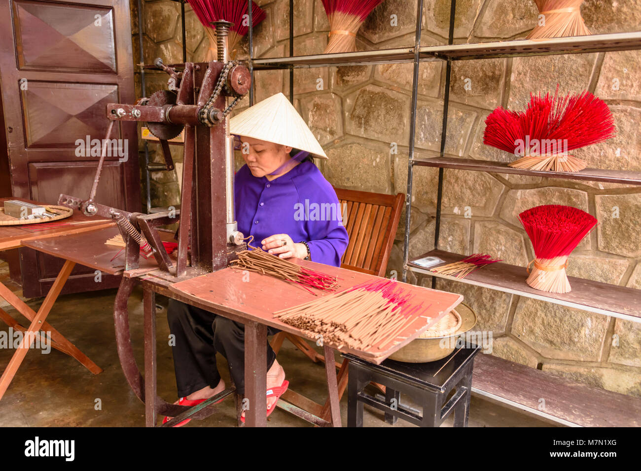 Un vietnamita donna che indossa un tradizionale di bambù cappello conico rende i bastoncini di incenso usando una vecchia macchina. Foto Stock