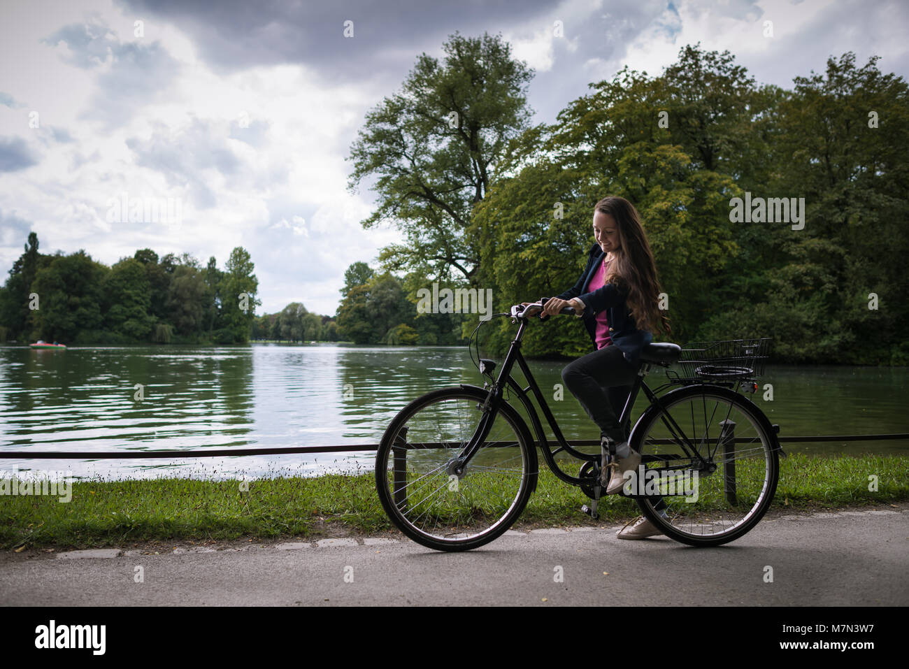 Giovane donna in bicicletta nel parco sullo sfondo di alberi e il lago. Attivo alla moda ragazza trascorre del tempo sulla natura. Vista laterale. Foto Stock