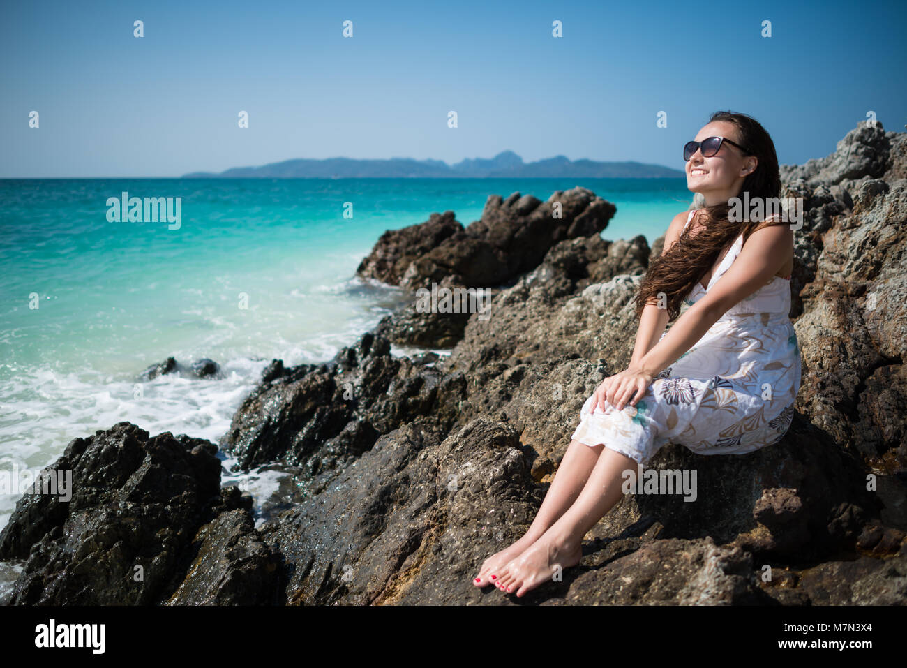 Giovane donna in abito siede sulla roccia sulla costa dell'oceano. Sorridente ragazza è godendo soleggiata giornata estiva. Tropical tavel destinazione Foto Stock