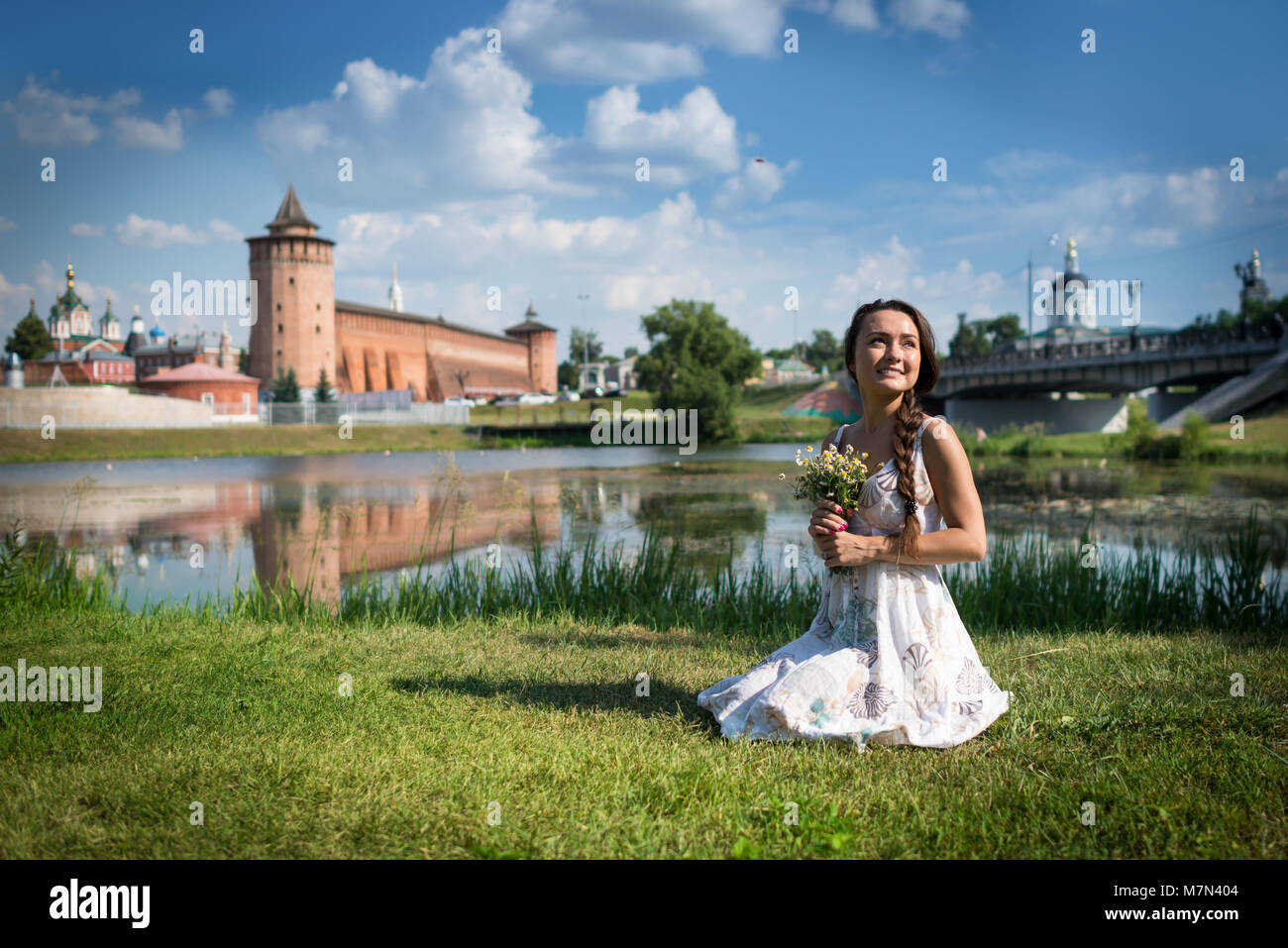 Giovane Donna con fiori in abito siede su erba vicino il Cremlino e la chiesa in una piccola città in Russia. Bella ragazza sulla riva del fiume Foto Stock