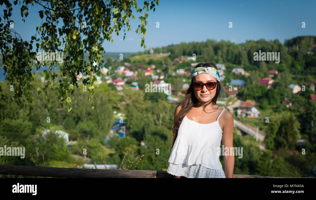 Giovane donna in sunglass e abito bianco sorge su di una collina nel giorno di estate e cercando nella fotocamera. Elegante ragazza nel parco vicino al piccolo villaggio di verde e di alberi Foto Stock