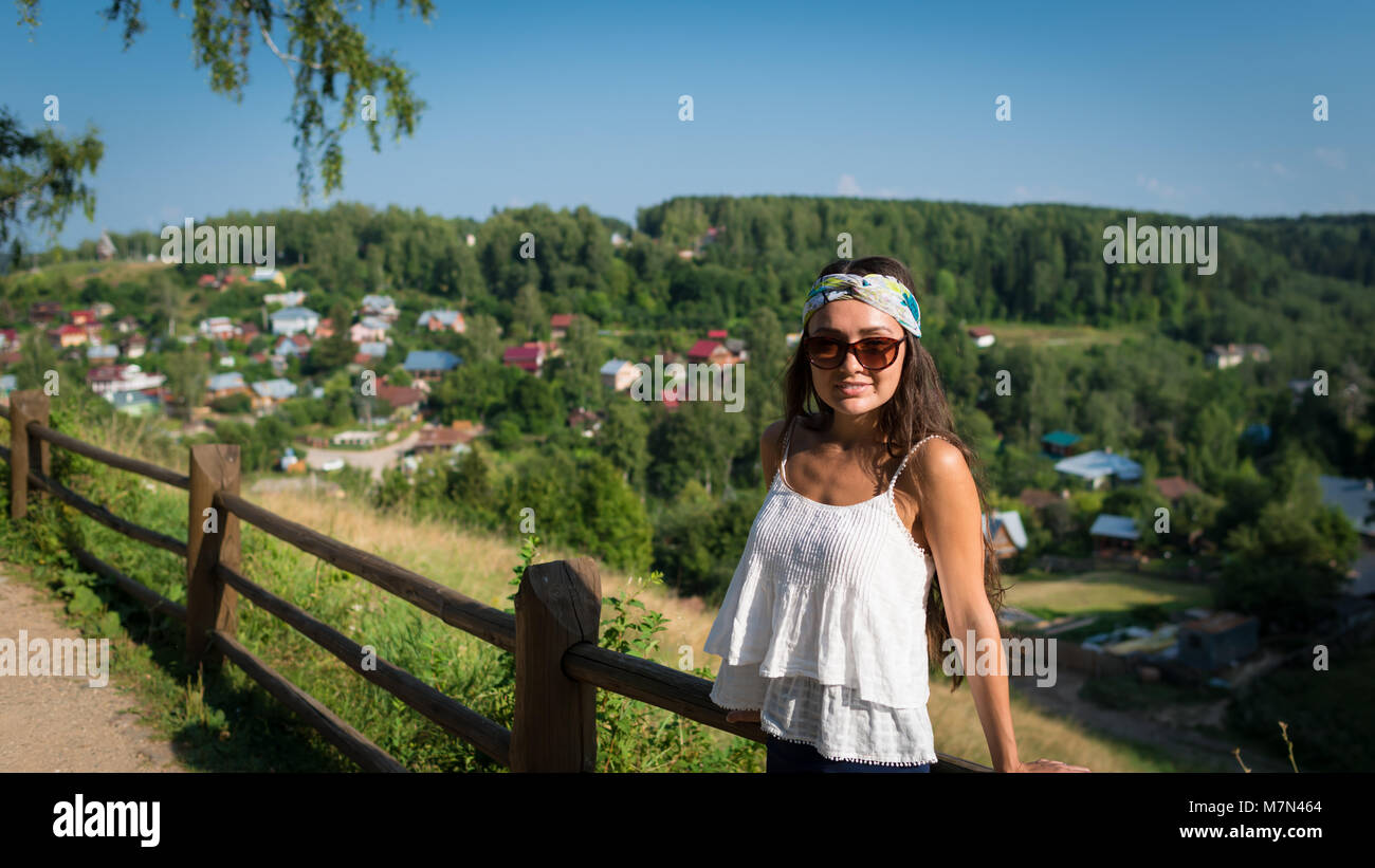 Giovane donna in occhiali da sole di bendaggio e sorge vicino al recinto sulla collina nel giorno d'estate. Ragazza in piedi nel parco su sfondo del piccolo villaggio e alberi verdi Foto Stock