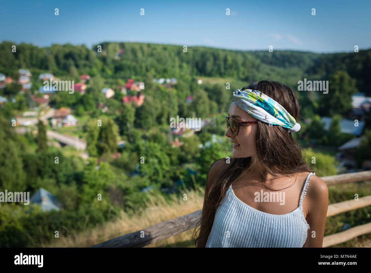 Vista laterale della giovane donna in abito bianco e la benda sulla testa permanente sulla collina nel giorno di estate nel parco vicino al piccolo villaggio e gli alberi in Russia Foto Stock