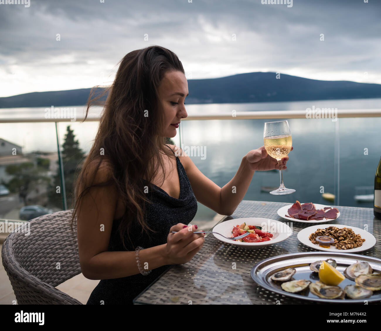 Giovani pensieroso donna siede sul balcone a tavola con ostriche, carne e insalata e detiene un bicchiere di spumante. Ragazza è pronto a mangiare. Mare e monti Foto Stock
