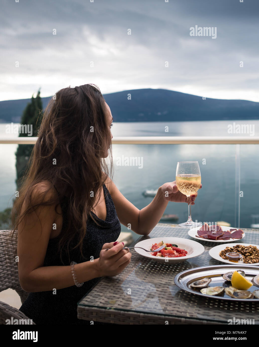 Giovane donna siede sul balcone a tavola con ostriche, carne e insalata e detiene un bicchiere di spumante. Ragazza guardando il mare e le montagne. Vista laterale Foto Stock
