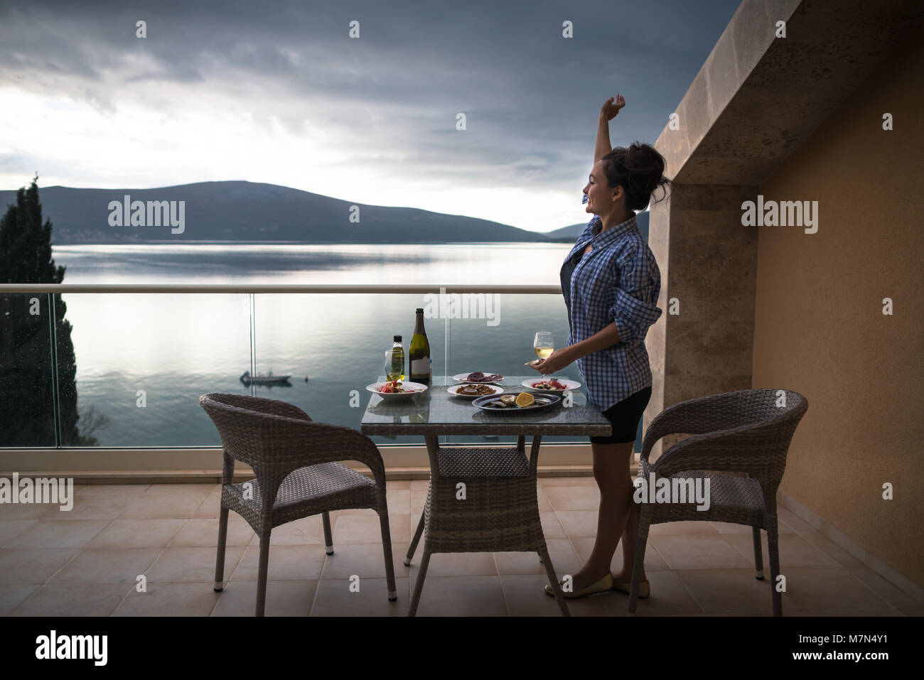 Giovane donna in piedi sul balcone dell'appartamento vicino a tavola con cibo e bevande e agitando a qualcuno. Ragazza sorridente con bicchiere di vino guardando il mare Foto Stock