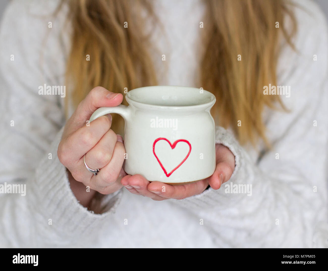 Una sezione intermedia di una bionda ragazza dai capelli tenendo una calda tazza di caffè con un amore cuore su indossando una calda felpa lavorata a maglia in un accogliente stile di vita shot Foto Stock