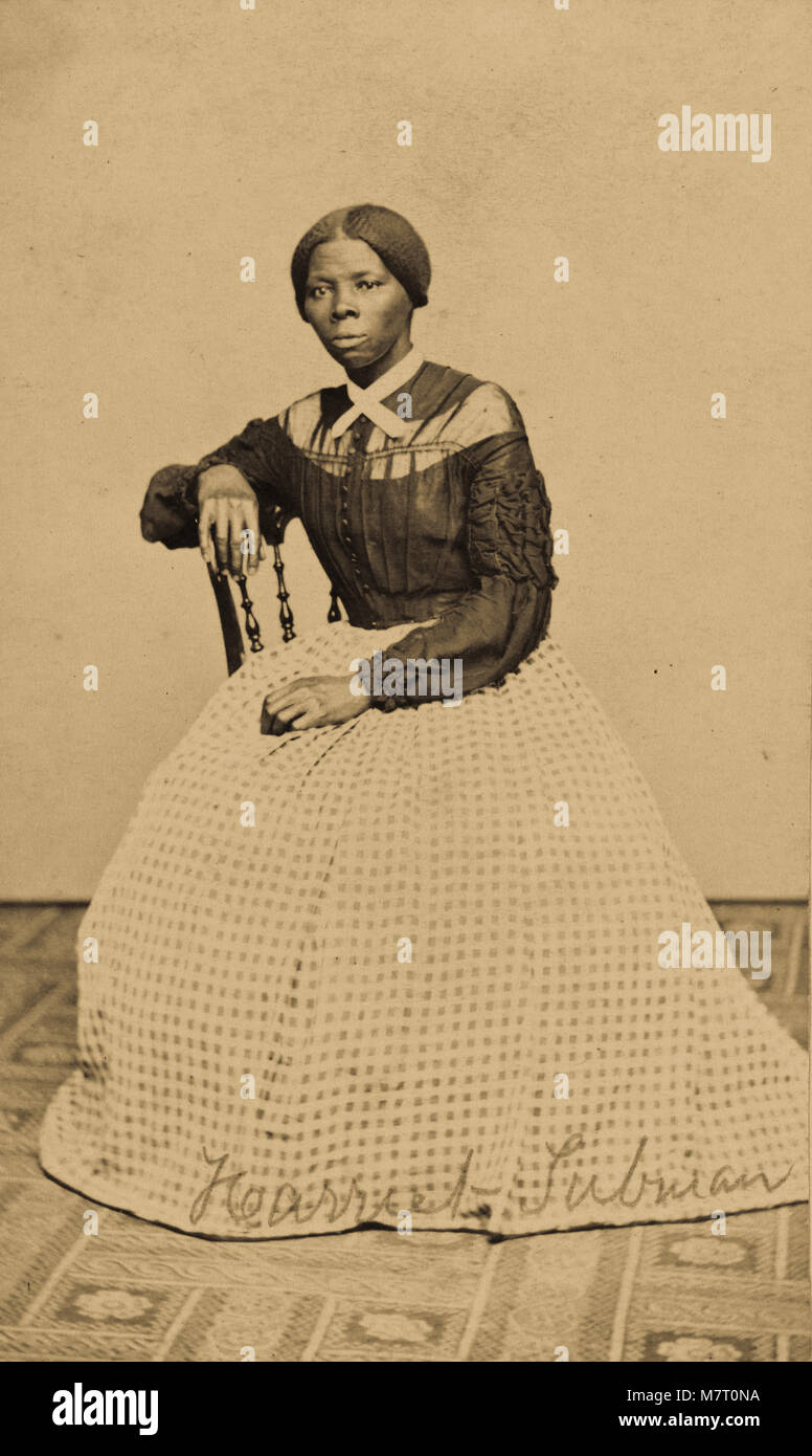 Fotografia mostra Harriet Tubman Ross (1820?-1913) alloggiata in una camera interna rivolta verso sinistra. Una mano appoggiata sul retro di una sedia in legno, un altro poggia in grembo. Una moquette a motivi geometrici ricopre il pavimento e la parete o caduta dietro di lei è un fustellato di colore chiaro. Tubman indossa un corpetto scuro che i pulsanti al centro anteriore e ha lasciato cadere manicotti con ruching pesanti e arruffata dettagli su le maniche. Vi è un pannello di tessuto più leggero attorno al giogo, con collo superiore lo stesso colore scuro come il corpo del corpetto. Un collare in pizzo con breve tails è attraversato e fissato tramite perni alla parte anteriore del suo collo. Foto Stock
