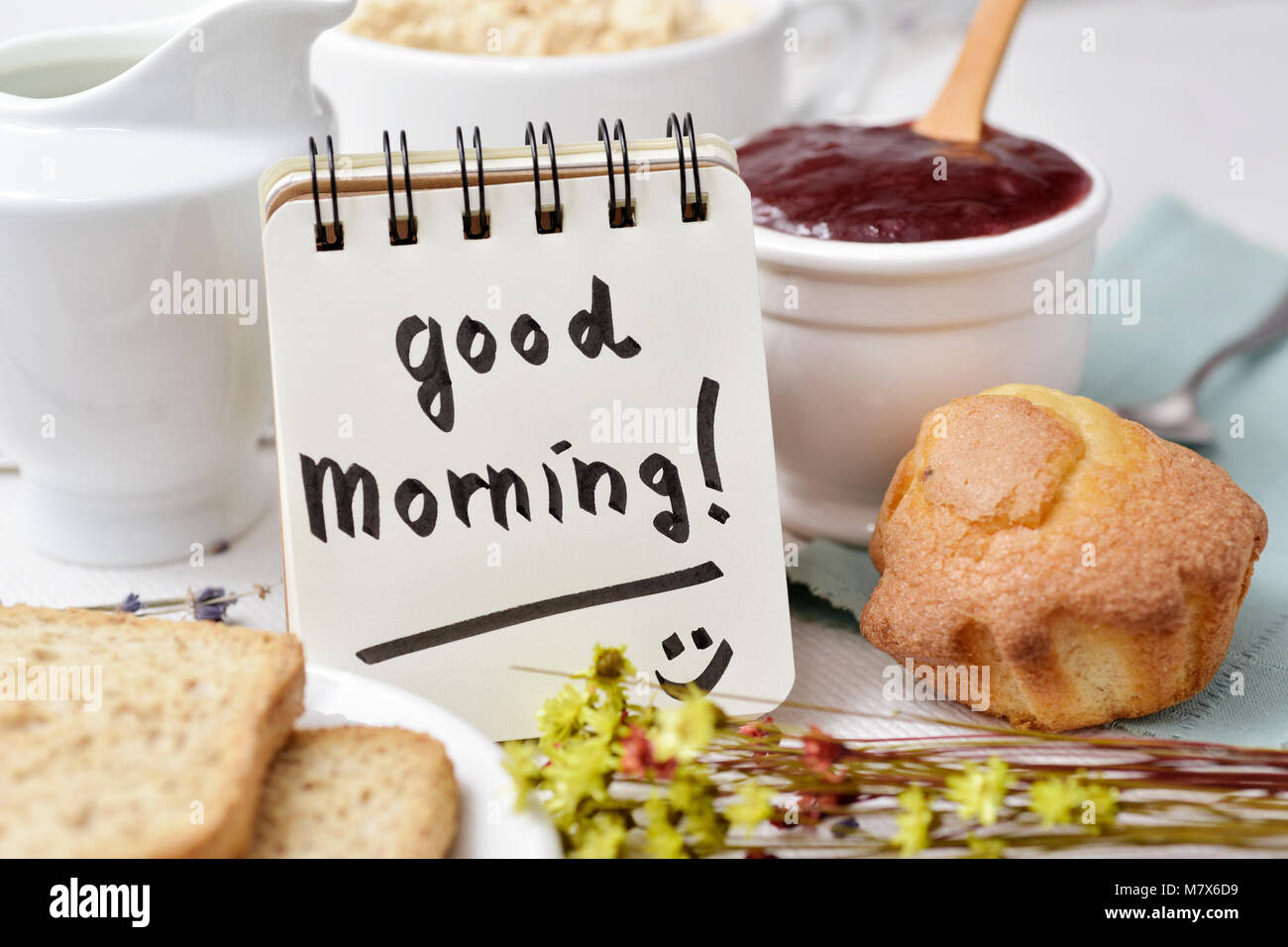 Primo piano di un tavolo per la prima colazione con un vaso di porcellana con cereali, qualche toast, una ciotola con marmellata e una nota con il testo di buona mattina scritto in Foto Stock