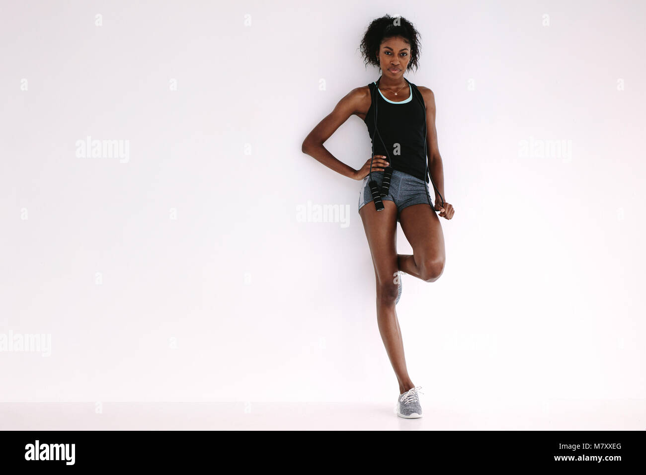 Montare e sano donna africana in piedi da un muro bianco con corda da salto. A piena lunghezza shot di sportive riposo dopo l'allenamento. Foto Stock